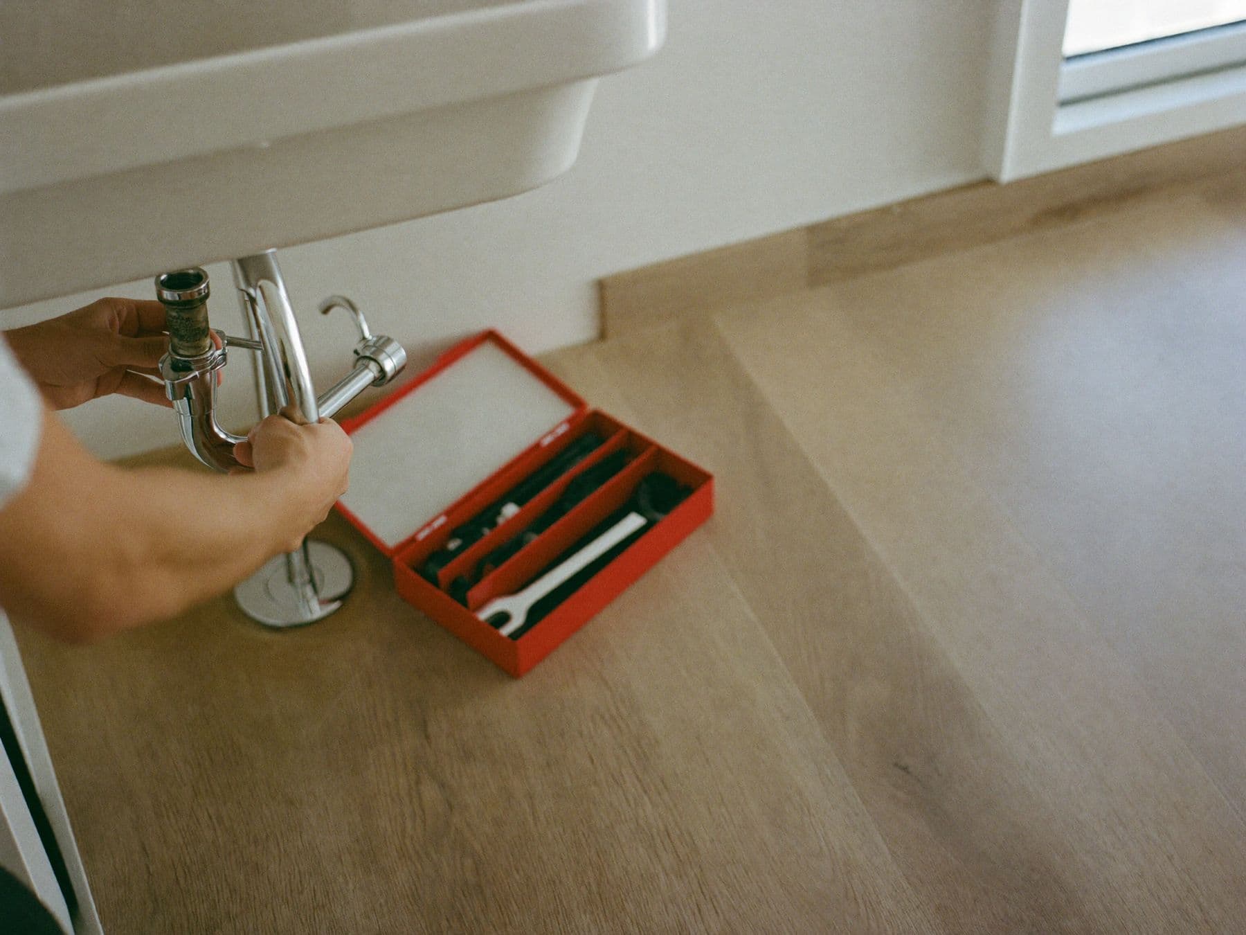 A plumber's hands fixing a sink pipe next to a red toolbox